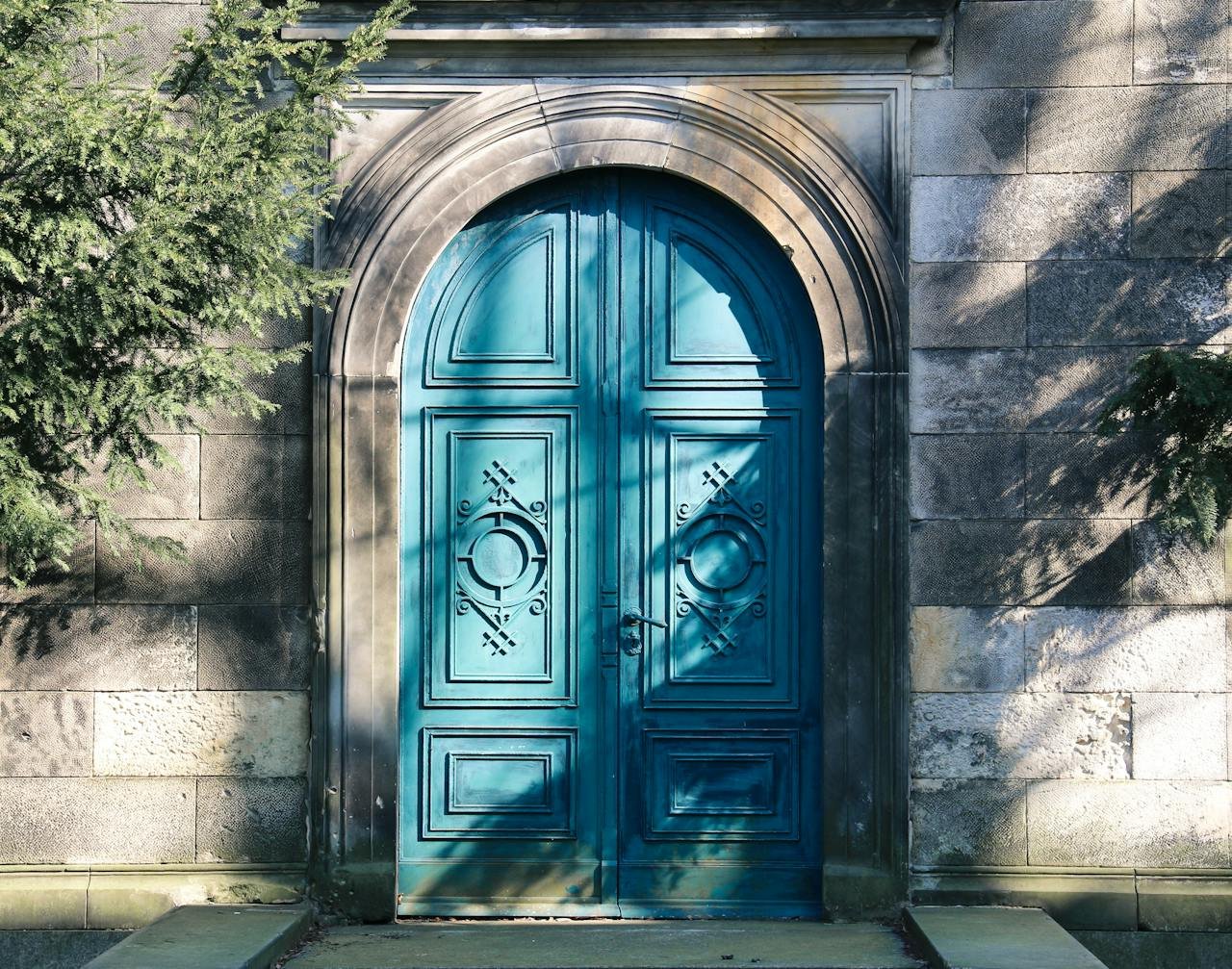 hero-img-01 A vibrant blue arched door with gothic designs. Shadows of trees add depth.